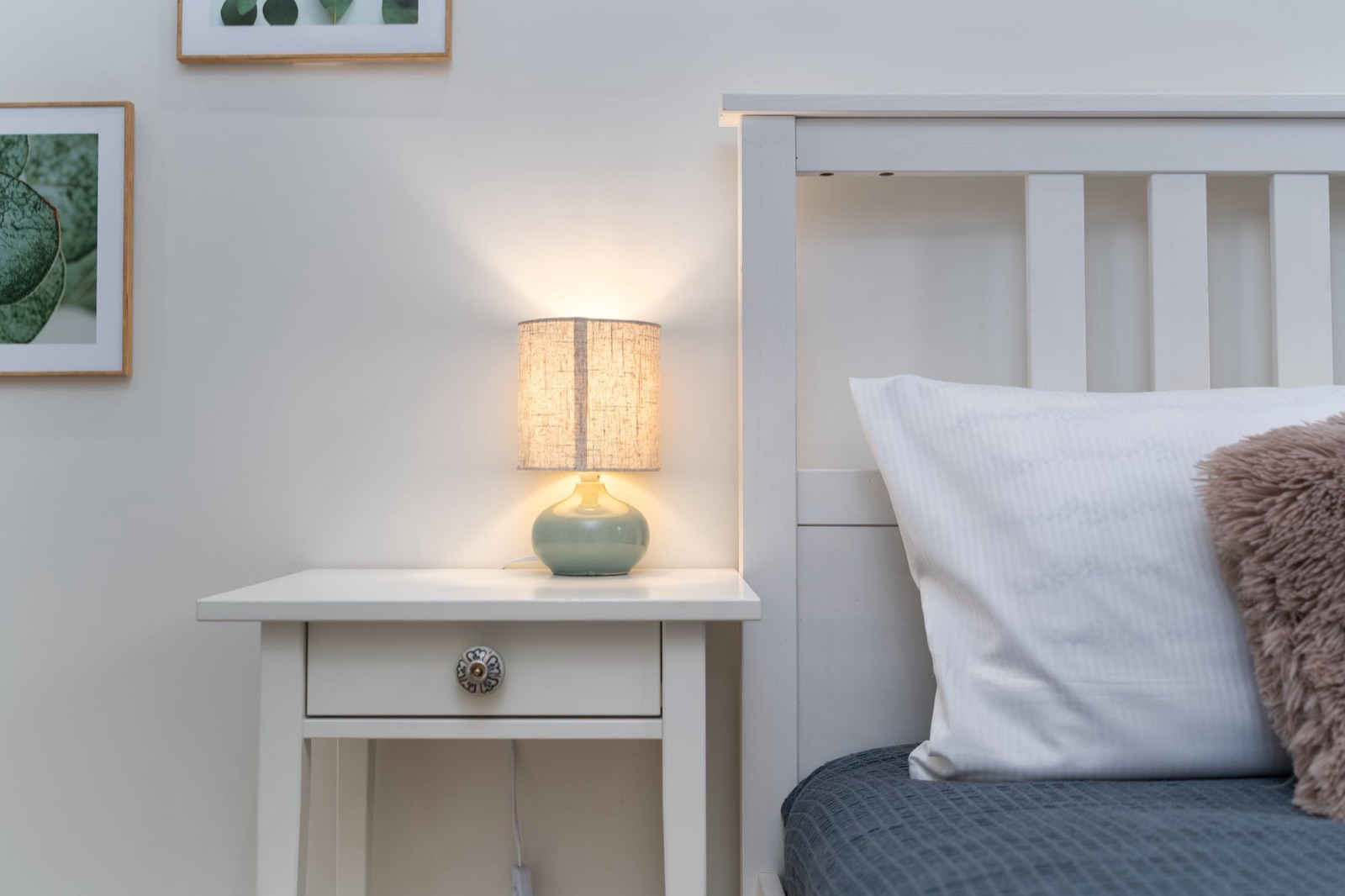 A small white bedside table with a glowing lamp beside a white bed and a gray throw.