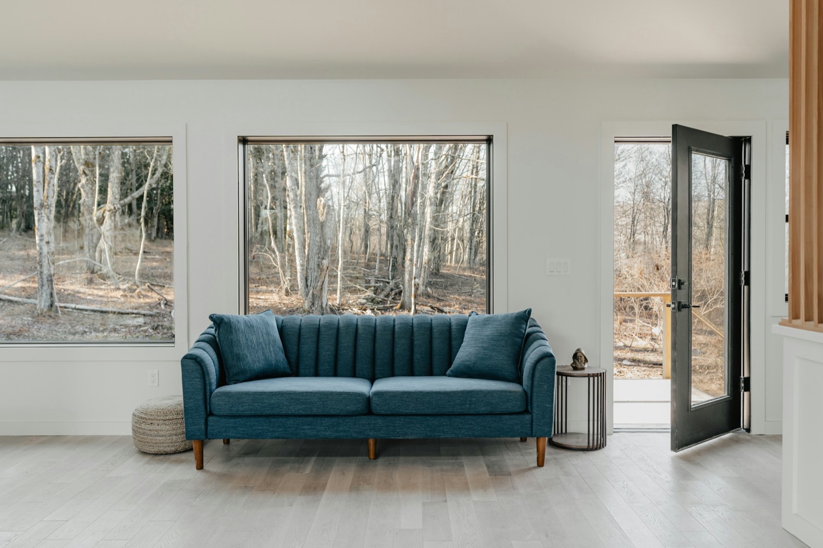 A blue channel-tufted sofa on tapered legs in a bright white room with tall windows onto bare trees.